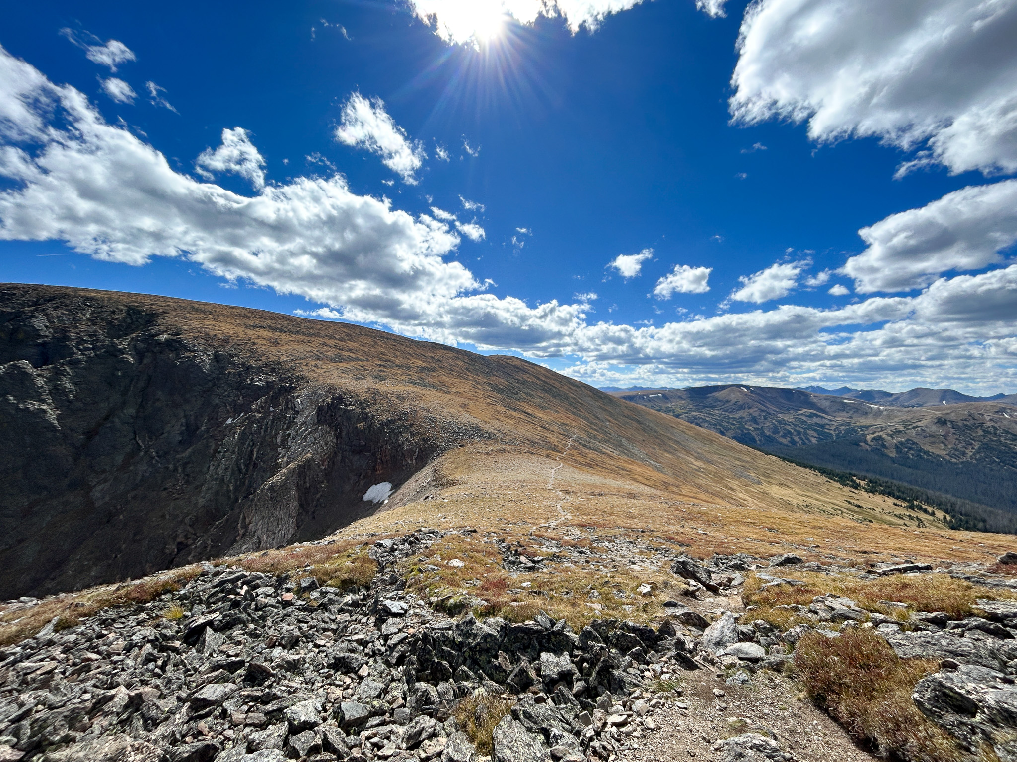 A scenic mountain landscape in Rocky Mountain National Park under a vast blue sky dotted with fluffy white clouds. The sun is visible on the upper right corner, casting a bright glare. A rugged mountain slope occupies the foreground, consisting mostly of rocky terrain with sparse vegetation, leading up to a ridge line that meets the sky. In the middle distance, the mountains continue to roll with varying shades of brown and hints of green, indicating sparse grasses or low bushes. In the far distance, a series of mountain ranges extend to the horizon, suggesting a vast wilderness area. It's a clear day, and the overall feel is of open space and natural beauty typical of a high-altitude environment.